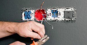 Close-up of an electrician's hands using pliers to strip wires during the installation of electrical outlets and switches in a wall. The image shows exposed wires and multiple socket frames against a gray background.